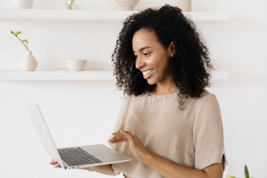A happy woman with curly hair standing and smiling at the laptop she’s holding