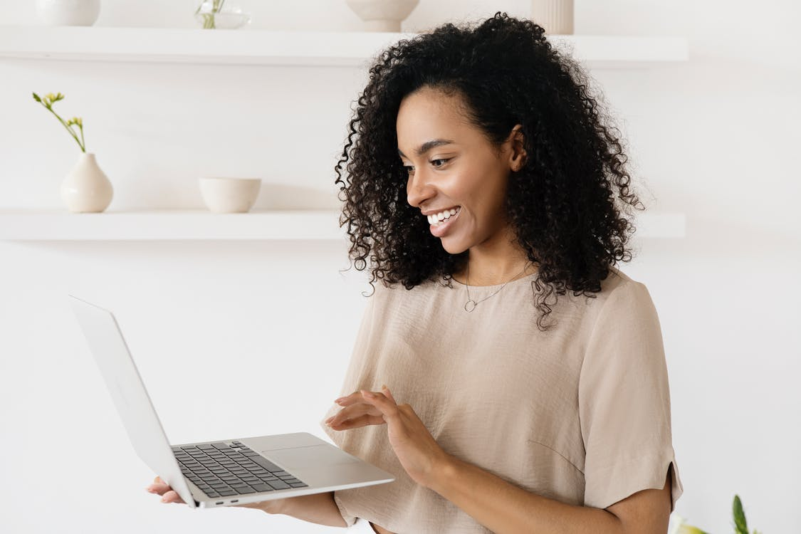 A happy woman with curly hair standing and smiling at the laptop she’s holding