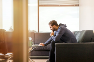 Man sitting on a couch and leaning over to use the laptop on the table in front of him