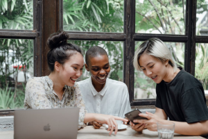 Three women sitting at a table smiling and pointing at a phone