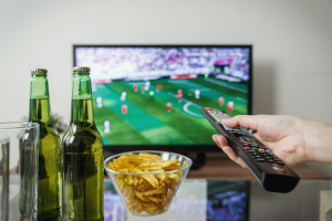 A person holding a TV remote with two bottles and a bowl of chips on a table in front of a TV screen displaying sports