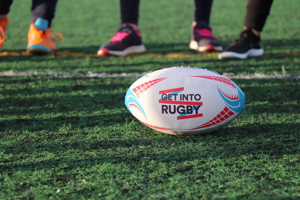 A rugby ball reading “Get into Rugby” on a field with players standing in the background