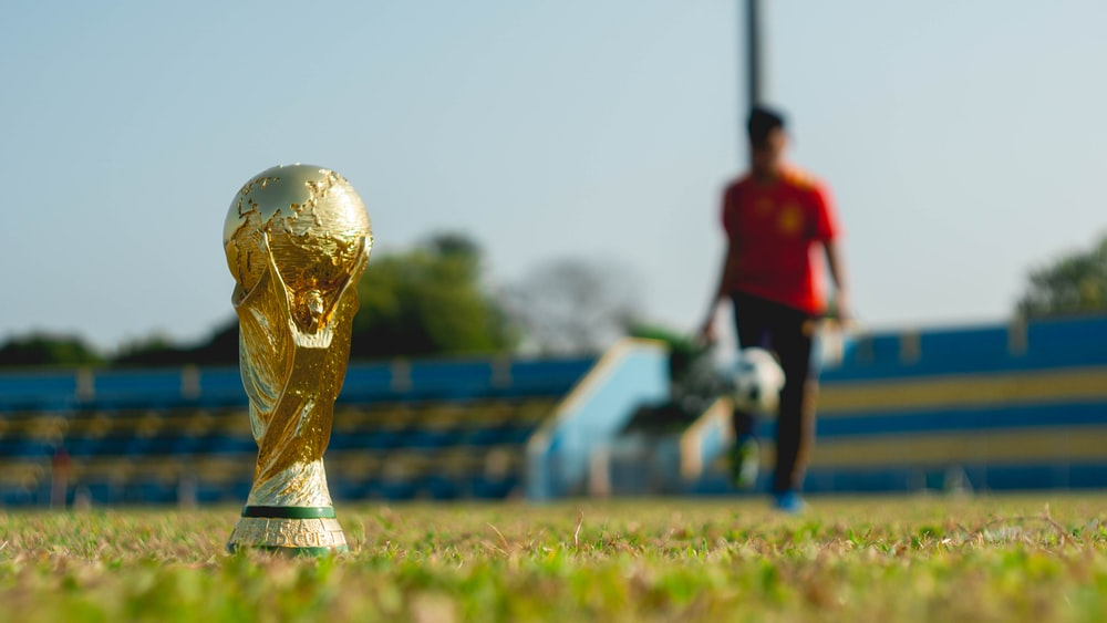 A FIFA World Cup trophy on the grass with a footballer practicing in the background